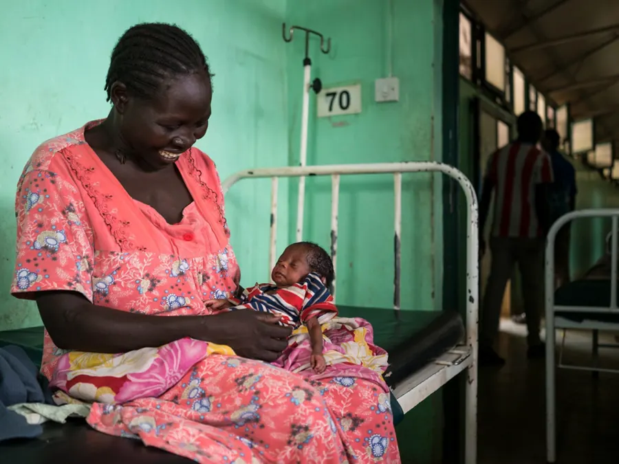 A mother sits on a ward bed holding her newborn baby, wrapped in a striped blanket