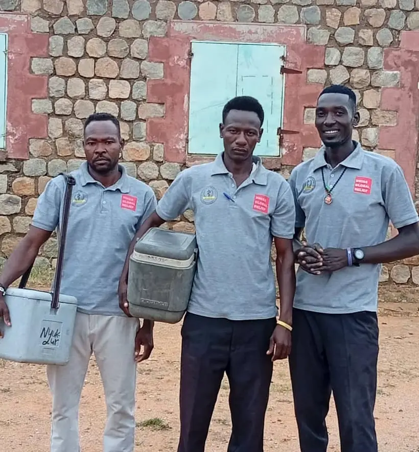 Three outreach health workers carrying vaccine cold boxes outside a stone building